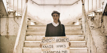 Μία γυναίκα κρατά μια επιγραφή που γράφει "Ζήτω ο Βασιλεύς. Καλώς ήλθες Μαννούλα μας"